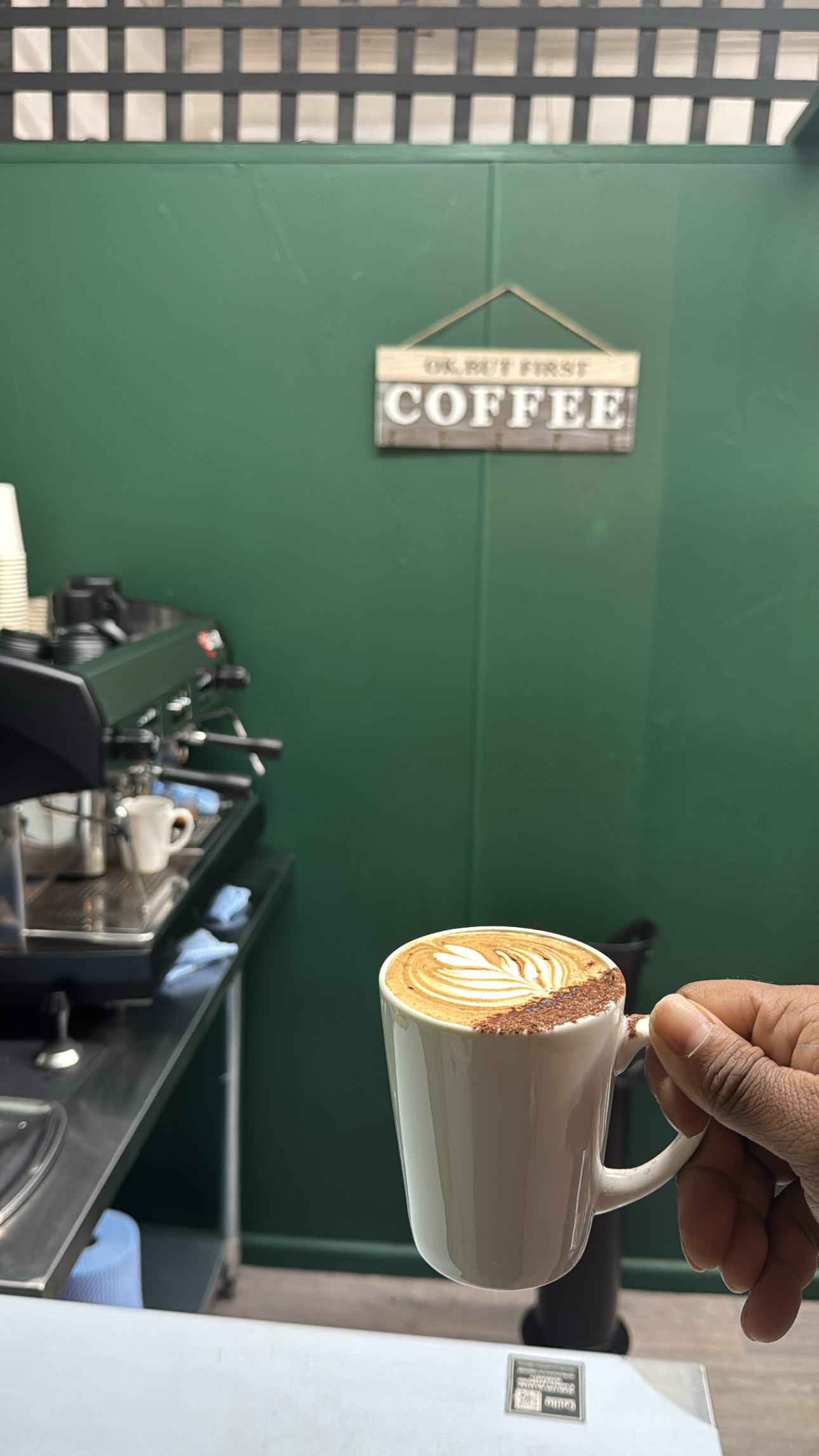 A latte with leaf latte art held up in front of the green wall and the OK But First Coffee sign