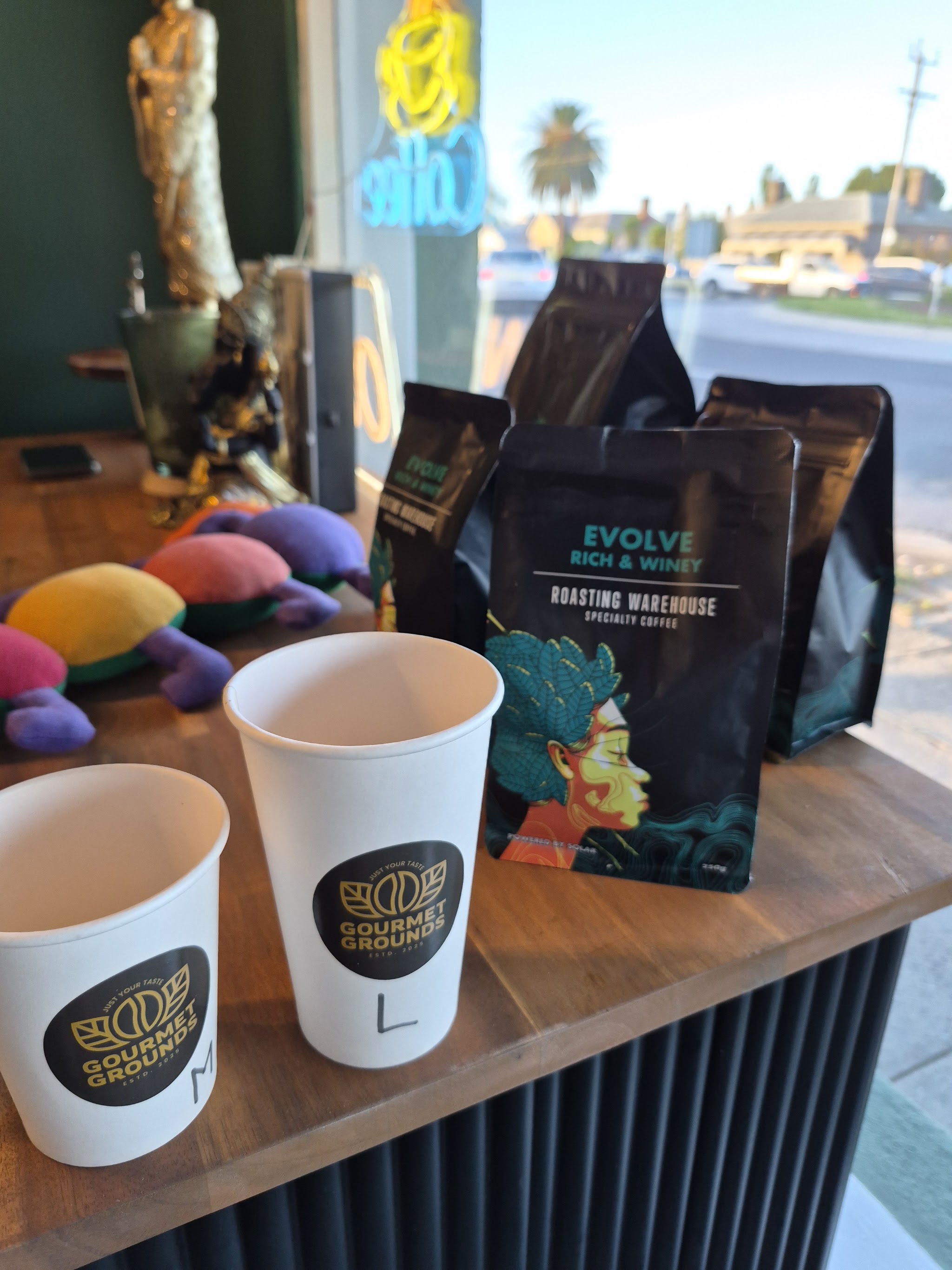 Two Gourmet Grounds takeaway cups beside bags of Roasting Warehouse Evolve coffee by the window